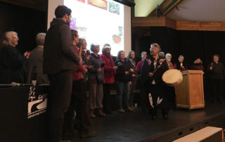 A group of people stand on stage with rattles while First Nations Elder Lolly Good drums and sings.