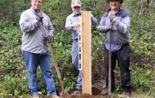 Three people stand by a post that has been put in a posthole; the middle person is ensuring that it is level.