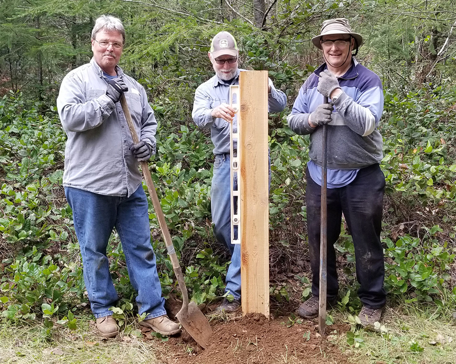 Three people stand by a post that has been put in a posthole; the middle person is ensuring that it is level.