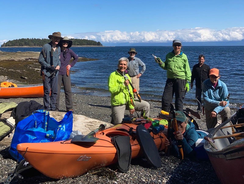 Group of people with gardening loppers, kayaks and canoes standing on pebble beach.
