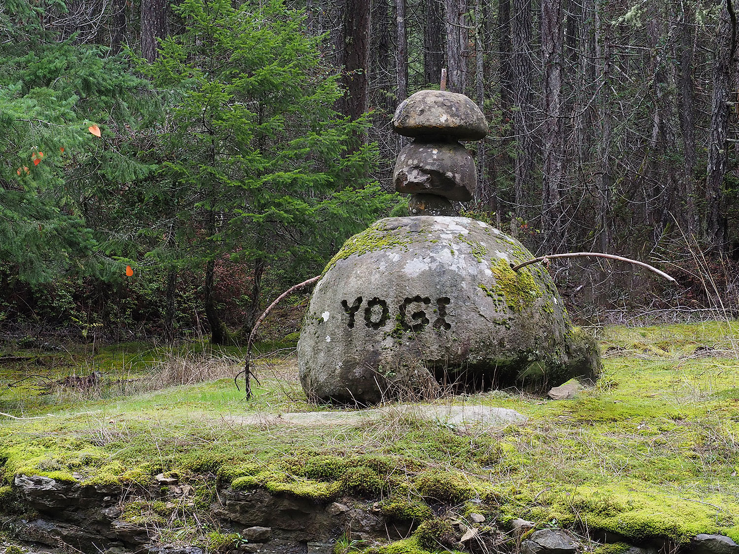 A sandstone sculpture by the edge of the road. It consists of a large round boulder with the word YOGI drilled into it and smaller stones piled on top. Two branches come out from either side. The overal effect is something like a human figure.