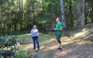 A man and woman standing in forest; the man is holding a binder of documents