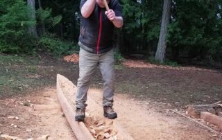 A man with an adze chops a cedar log into the rough shape of a canoe