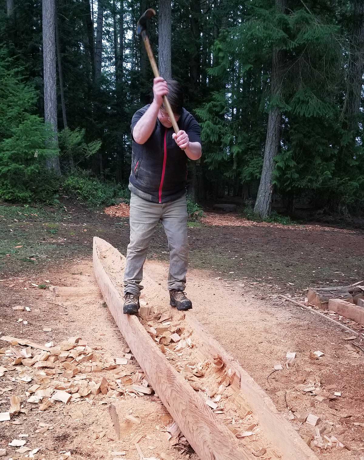 A man with an adze chops a cedar log into the rough shape of a canoe