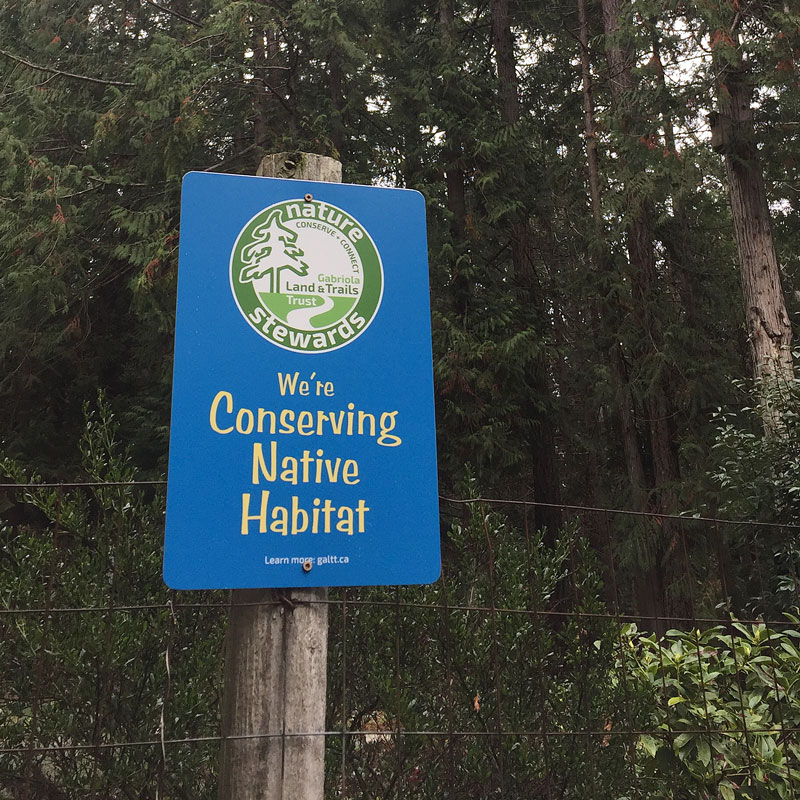 A sign on a fence in front of forest says, "We're conserving native habitat."