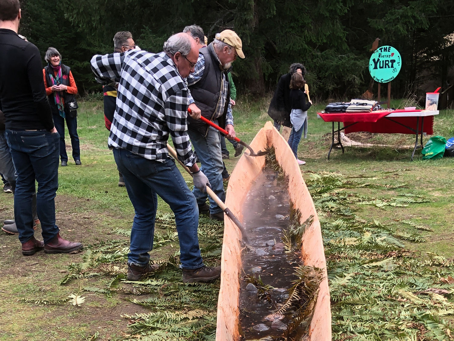 Two men use shovels to remove rocks from hot water in a traditionally crafted cedar canoe, as part of the process of using steam to spread the sides of the canoe into its desired shape.