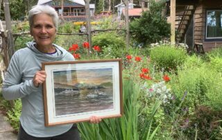 A woman stands in a garden holding a framed painting of ferries at sea.