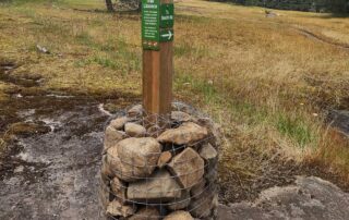 A signpost stands at the edge of a moss meadow. The base is set in a stack of heavy rocks, held together with wire fencing.