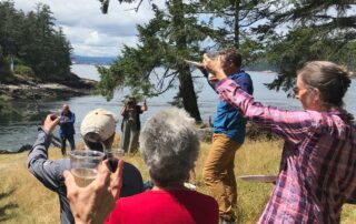 A group of people raise a glass of wine to toast Jack Schick at his celebration of life. The setting is a park near the ocean.