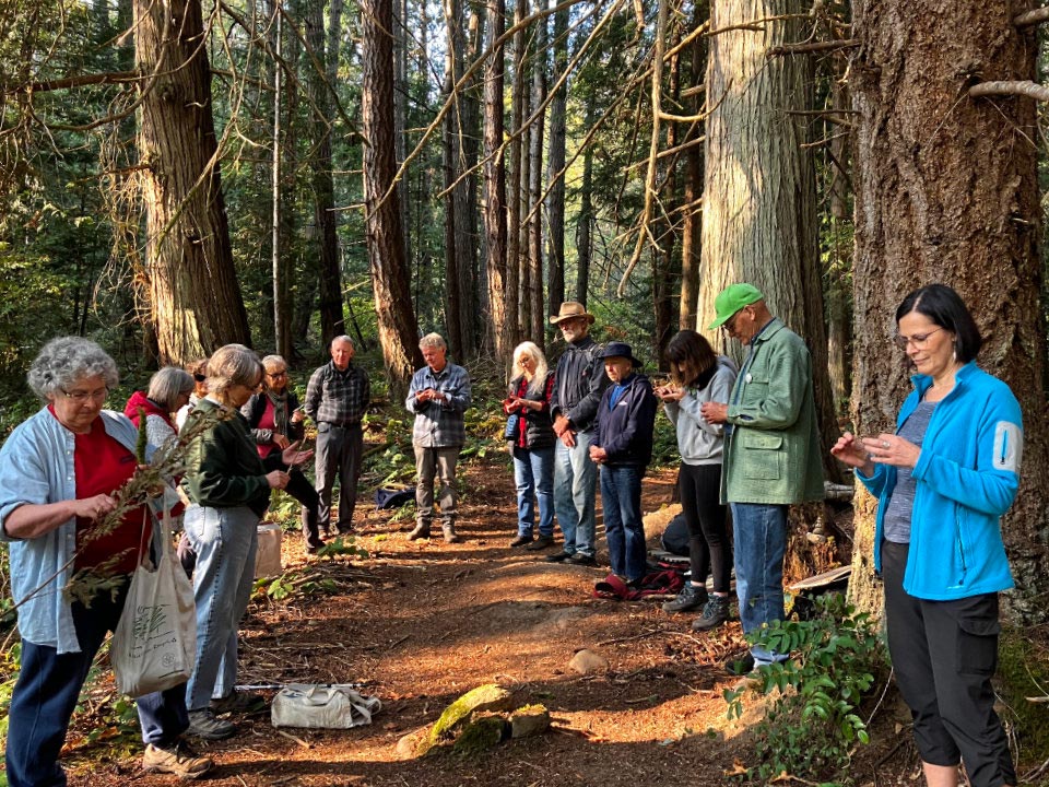 A group of people stand in a circle on a forest trail. Most are holding a natural object and examining it with close attention.