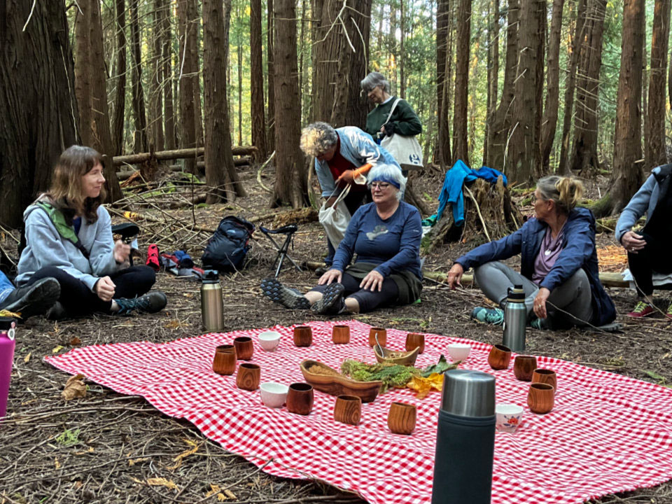 A group of people sit around a red and white checked tablecloth in a forest clearing. On the cloth are wooden cups and other utensils.