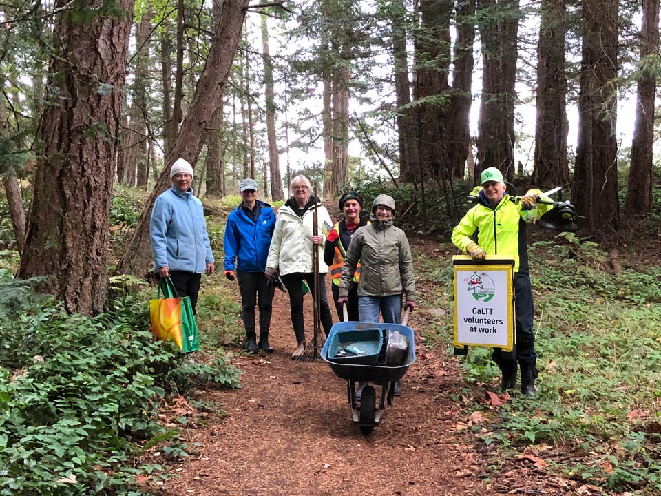 A group of people on a park trail. Several hold tools, one pushes a wheelbarrow, and another carries a sign that says, "GaLTT volunteers at work."