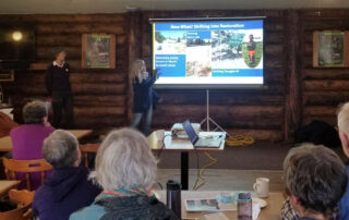 A group of people sit listening to two women giving a slideshow presentation.