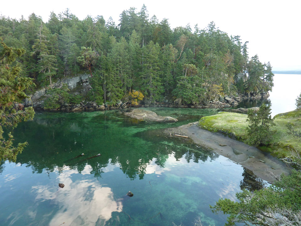 A protected bay with a very narrow channel between two islands. The water is blue and emerald green and the rocky shores are covered with new green growth.