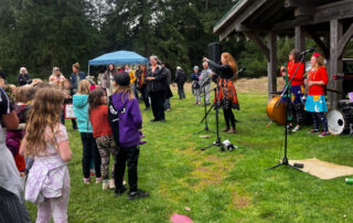 An audience of children and adults listens to a speaker at an Earth Day presentation. Members of the Kerplunks band stand in the background, waiting to perform.