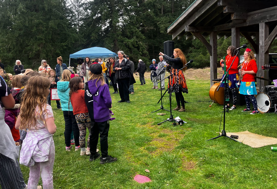 An audience of children and adults listens to a speaker at an Earth Day presentation. Members of the Kerplunks band stand in the background, waiting to perform.