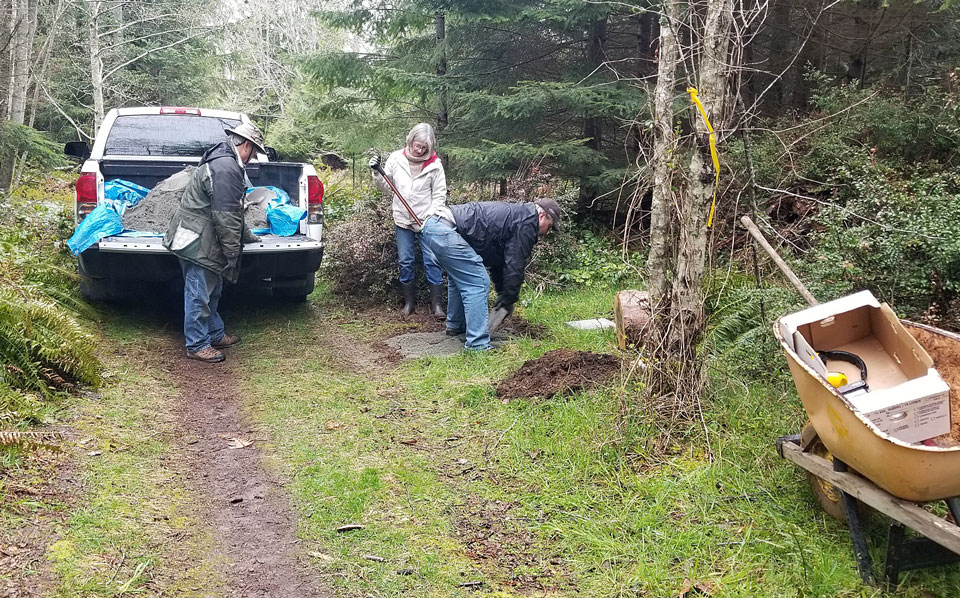 The photo shows a workparty on a trail. A man and a woman with a rake work on a gravelled area just off the trail, and another man stands behind a pickup truck; its tailgate is down, showing a large pile of fine gravel. There is a wheelbarrow and more tools in the foreground.