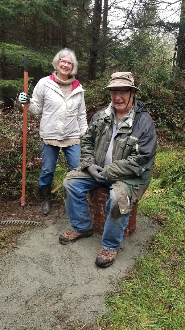 A woman with a rake stands next to a man sitting on a stump. Both are smiling.