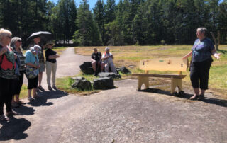A woman stands by a beautifully crafted wooden bench, speaking to a group of onlookers.