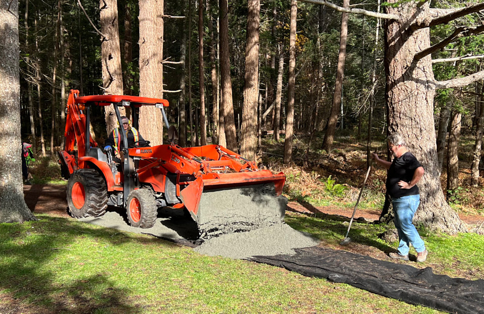 A man with a shovel stands watching a front-end loader spread gravel on a pathway liner in a park.