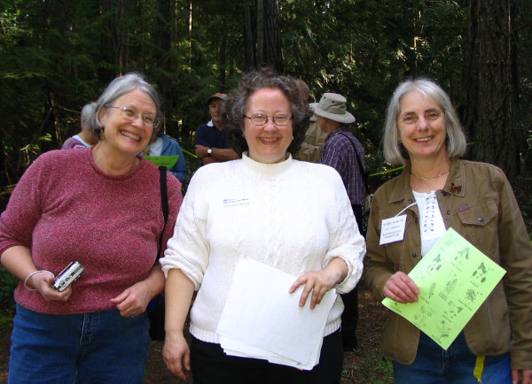 Three women stand smiling at an outdoor event.