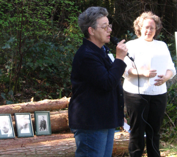 A woman with a microphone speaks at an outdoor event. Beside her another woman listens. Framed historical photos rest on piled logs behind her.