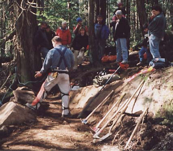 The photo shows a workparty on a trail. A group stands in the back listening to a man in working clothes talk. An assortment of pickaxes and shovels leans against a bank in the foreground.