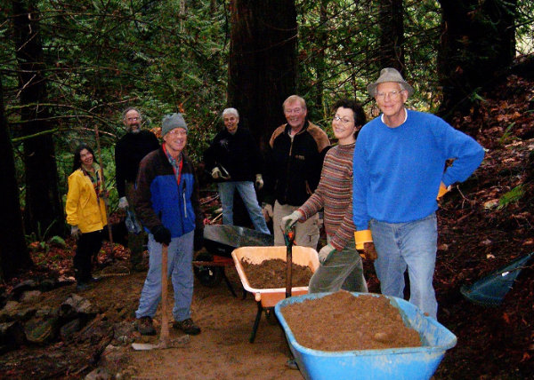 A group of men and women with tools stand on a trail beside wheelbarrows full of dirt.