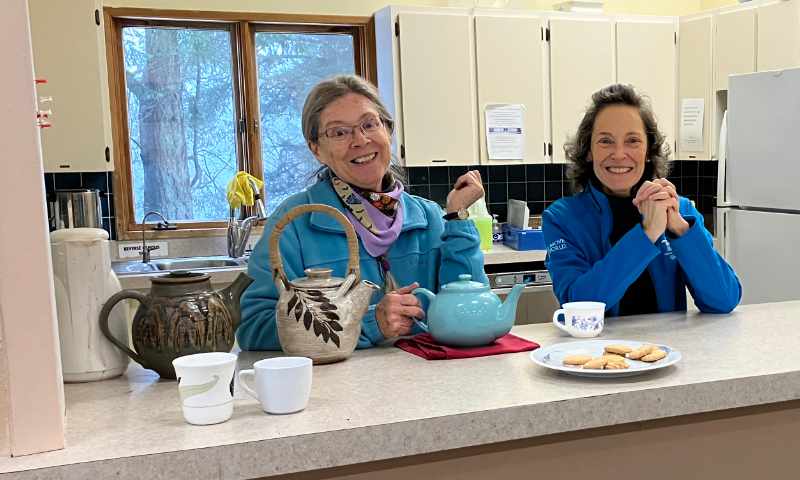 Two smiling women stand behind the food counter at the Gabriola Island Community Hall. A plate of cookies and teapots sit on the counter.