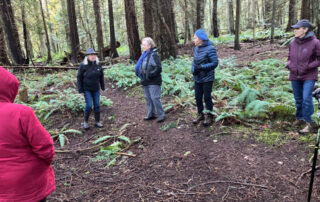 A photo of a group of people standing on a trail in a forest listening to a woman speak.