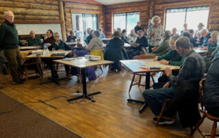 The photo shows a large number of people witting at tables in preparation for the start of a meeting.
