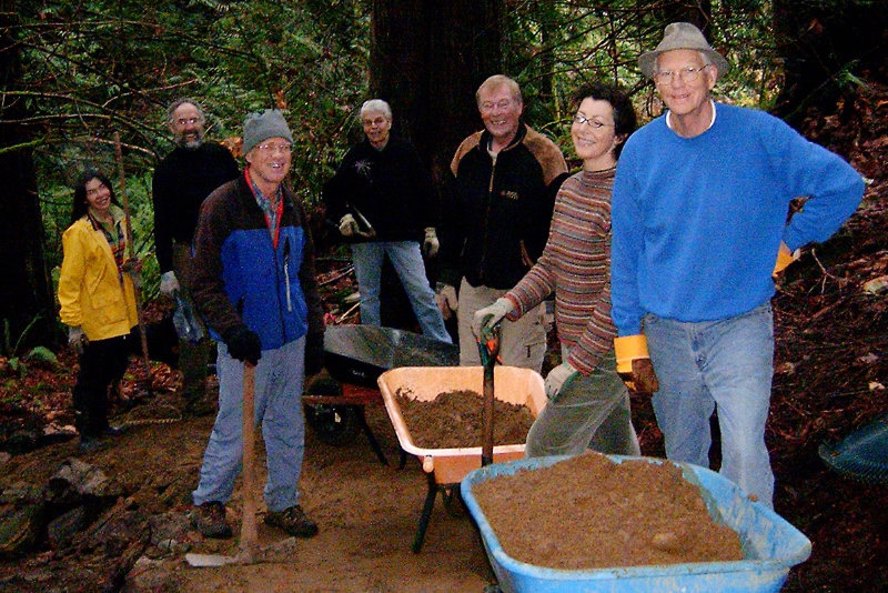 The photo shows a group of people with wheelbarrows full of dirt on a forested trail.