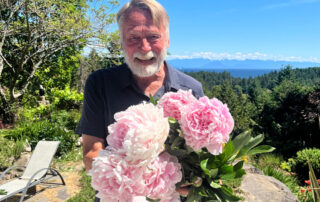 A man with a beard holds giant pink peonies, grinning with delight.