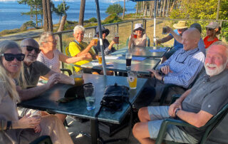 A group of smiling people with drinks sit around a table on a patio. Beyond them is the ocean.