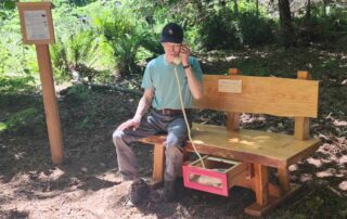 A man sits on a beautifully crafted resting bench in a park. A drawer under the bench holds a corded phone; it is open and he holds the phone to his ear.